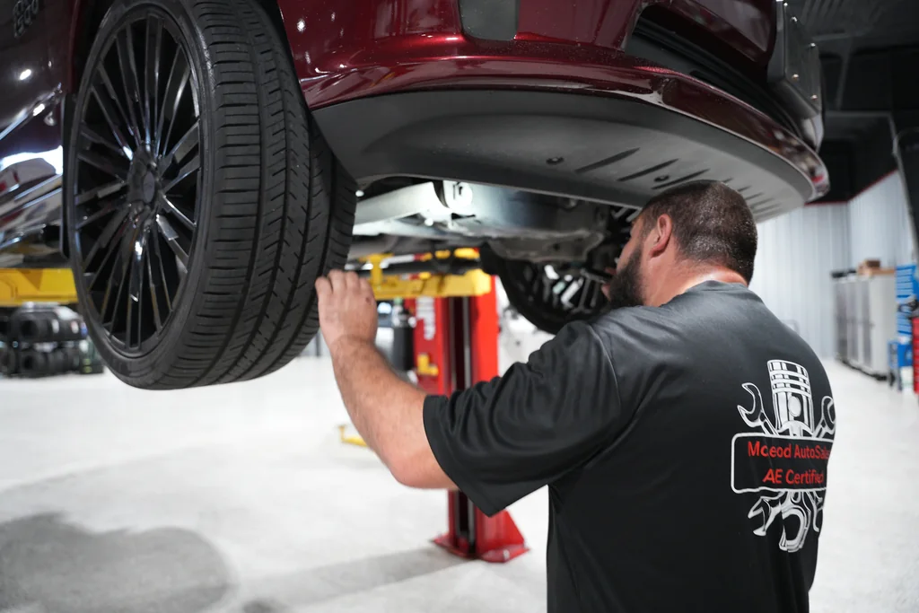 Técnico inspeccionando la parte inferior de un vehículo elevado junto a la rueda delantera.